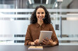 © Liubomir - Smiling woman wearing a headset and holding a digital tablet, sitting at a desk in a modern office, ready to provide customer support and online assistance