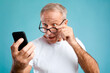 © Prostock-studio - A cheerful mature man in glasses expresses excitement while reading a smartphone screen. He celebrates good news in a bright blue studio background. His surprise and joy are clearly visible.