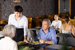 © JackF - Polite young waiter serving fresh vegetable salads to guests, positive elderly couple, visiting restaurant for dinner, sitting at table with glasses of wine ..