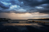 Dramatic coastal landscape with dark storm clouds, sunset light and wet asphalt reflecting the sky. Moody seascape with harbor elements, calm water and a distant shoreline under heavy weather.