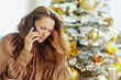 © Alliance - A middle-aged woman on a phone call appears deeply worried, touching her temple with a distressed expression against a blurred, festive Christmas tree background.