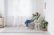 © Pixel-Shot - Young woman sitting in armchair near light curtain at home