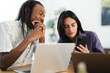 © Studio Marmellata - A diverse couple shares a moment, looking at a smartphone together. They are seated at a table with laptops, engaged in a digital interaction. The man smiles.