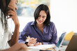 © Studio Marmellata - Two diverse individuals collaborate at a desk. A smiling woman in a purple shirt writes in a notebook with a pencil. Her colleague is beside her. They are focused on their work.