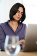 © Studio Marmellata - A focused young woman with dark hair works diligently on her laptop. She is wearing a purple shirt, concentrating on her task in a bright office setting. A glass of water sits nearby.