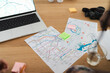 © Studio Marmellata - People plan a trip using a laptop, physical maps, and sticky notes on a wooden table. A camera and notebook are also present, indicating travel preparation.