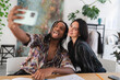 © Studio Marmellata - A diverse young couple takes a fun selfie indoors. The man with dreadlocks makes a silly face, sticking out his tongue, while the woman smiles happily beside him. They capture a joyful moment.