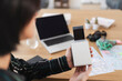 © Studio Marmellata - A person holds a compact photo printer, watching as a freshly printed picture emerges from the device. A laptop and travel maps are visible on the wooden desk.