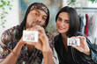 © Studio Marmellata - A happy diverse couple smiles at the camera, each holding up a small instant photo. The man holds his picture, and the woman holds hers, sharing a joyful moment.