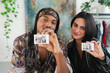 © Studio Marmellata - A diverse couple smiles, holding up instant photos of themselves. They share a joyful moment indoors, surrounded by plants and a painting.