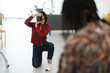 © Studio Marmellata - A smiling young female photographer kneels, holding a camera to her eye, capturing a shot of a person in a bright indoor studio setting. She is focused on her work.
