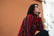 © Studio Marmellata - Pensive young woman with dark hair gazes intently upwards, hand resting on her chin. She wears a striped red and black shirt against a plain brown studio background.