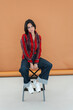 © Studio Marmellata - A stylish young woman with dark hair sits confidently on a stool, gazing directly at the camera. She wears a striped shirt, wide-leg jeans, and white athletic shoes in a studio.