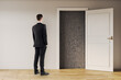 © Who is Danny - Businessman standing in front of an open door revealing a dark textured wall, symbolizing uncertainty and decision making in a light interior background.
