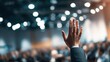 © Sergey - close up of a raised hand of a business professional during a modern conference, blurred audience and stage lights behind, clean corporate setting