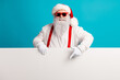 © deagreez - Santa Claus with red suspenders and a fluffy beard smiles behind a blank banner on a bright blue studio backdrop ready for Christmas promotion