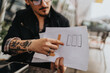 © qunica.com - Close-up of a man holding papers with sketches, presenting ideas in an outdoor setting. Details include hand tattoos, stylish accessories, and a focus on collaboration and creativity.