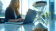 © Ruslan - An hourglass stands on a table next to a woman working on a laptop – a concept for a deadline or getting work done quickly