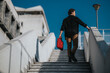 © qunica.com - A man in casual business attire climbs a concrete stairway outdoors, carrying a red bag. The urban setting features modern architecture and a bold blue sky, conveying travel, work, and momentum.