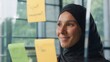 © Yuliia - Satisfied islamic business woman analyzing stickers on glass in office Muslim businesswoman in hijab company CEO entrepreneur female girl reading sticky notes data plans smiling management work task