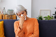© Xavier Lorenzo - Senior sick woman sitting on sofa and blowing her nose with a tissue, feeling sick with a cold or flu