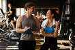 © Home-stock - Happy woman talking to her male trainer, holding bottle with water, having break during workout at modern gym interior
