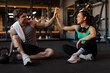 © Home-stock - Two gym-goers giving high five while sitting on the gym floor, man and woman relaxing after workout, surrounded by fitness equipment