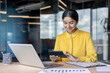 © Tetiana - Young smiling Indian woman working in the office, sitting at a desk with a laptop and documents, using a calculator