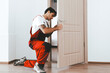 © Prostock-studio - A skilled worker kneels on the floor to fix a door lock in a contemporary room with wooden flooring and clean walls. The craftsman wears a red and white work outfit, focused on the task.