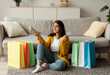 © Prostock-studio - A joyful arab woman sits on the floor among colorful shopping bags, holding a credit card and a new shoe while enjoying the experience of online shopping from her cozy home.