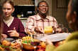 © DragonImages - Diverse group of young adults and middle aged friends sharing Thanksgiving dinner, Black man smiling while passing corn to Caucasian woman, food and wine on table, people interacting