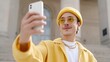 © Bella - Fashionable young Latin man in a bright yellow outfit, beanie, and round sunglasses takes a selfie with a white smartphone against a grand stone building.