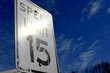 © Gregory - A reflective white 'SPEED LIMIT 15' sign angled against a dramatic, deep blue sky with wispy clouds, featuring a bright sun glare.