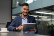 © Prostock-studio - A cheerful Middle Eastern CEO sits at a table in a modern office, smiling as he checks the latest business mobile application on his tablet. He is dressed in a nice suit, enjoying the moment.