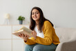 © Prostock-studio - A happy arab woman sits comfortably in her armchair with a book in hand. She is taking a break from work, smiling at the camera and enjoying her free time in a relaxed home setting.