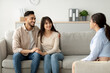 © Prostock-studio - A happy middle eastern couple sits on a couch, holding hands and smiling as they engage in a therapy session. A therapist listens attentively, creating a supportive environment for their discussion.