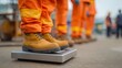 © Bussakon - Construction workers in orange high visibility gear standing on a metal digital scale at an outdoor work site
