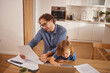 © Stockphotodirectors - A man reviews paperwork while talking on the phone, assisting a young child with a laptop. Their kitchen is bright, reflecting an enjoyable family time.