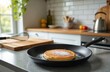 © Pete - Pancake in a frying pan on kitchen countertop. Cooked round dessert cake sprinkled powder sugar. Whisk, kettle, cutting board on background. Delicious breakfast food at home prepared bakery meal.