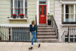 © Studio Marmellata - A young woman walks along a London street in Camden, passing traditional terraced houses with a vibrant red door and concrete steps. Her hair blows in the breeze.