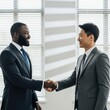 © Jesmin - Two businessmen of different ethnicities shaking hands in a modern office setting with natural light.