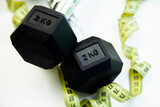 Black dumbbells resting on a clean white surface, with a yellow measuring tape nearby, representing fitness, exercise, and health aspirations in a workout environment