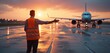 © Pete - Airfield specialist directs plane at sunset. Ground staff regulates aircraft traffic on runway. Airport worker in safety vest communicates with pilot using hand signals and controls movement.