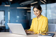 © Tetiana - A young Indian businesswoman in a yellow shirt sits at a desk in the office and works on a laptop with a smile