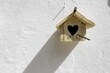 © Travel 'n' Lifestyle - View of a wooden birdhouse with a heart-shaped opening casts a long, stark shadow on a textured white wall, Ã“bidos, Leiria District, Portugal.