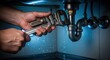 © Olga Ко - Close up of a plumber's hands using a wrench to fix a leaking metal pipe under a sink, water droplets falling, blue toned lighting, professional repair concept