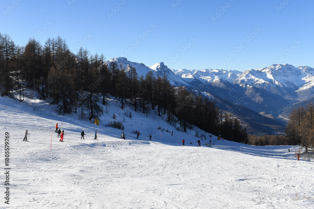 Skiers and snowboarders descending a ski slope in Sauze D'Oulx ski resort, Turin, Italy. Tree lined piste overlooking beautiful snowcapped mountains and the Milky Way ski area  in the Piedmont region.