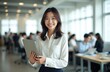 © Maryna - Young Japanese businesswoman smiles holding tablet computer. She stands in modern office with blurred team working behind her. Pro woman uses tech device for work.