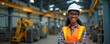 © Maryna - Smiling Black woman wears hard hat and safety vest in factory. She stands arms crossed near machinery, representing industrial work and safety protocols. Female engineer in building.
