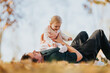 © qunica.com - A father lies on the grass in an autumn park, lifting his baby girl for a joyful moment among leaves and sunshine. Warm, tender family time outdoors.